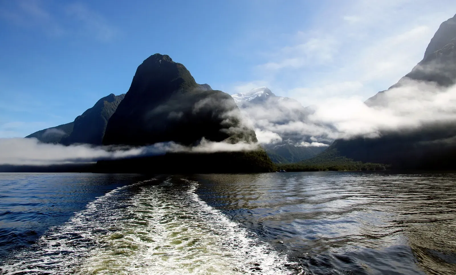 Boat wake crossing Milford Sound beneath mist-wrapped fjord peaks — Fiordland