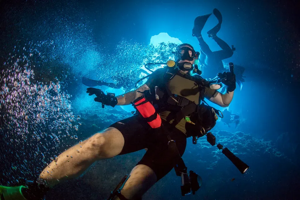 Two divers ascend trailing bubbles inside a bright blue rock chimney — Blue Hole, Guam