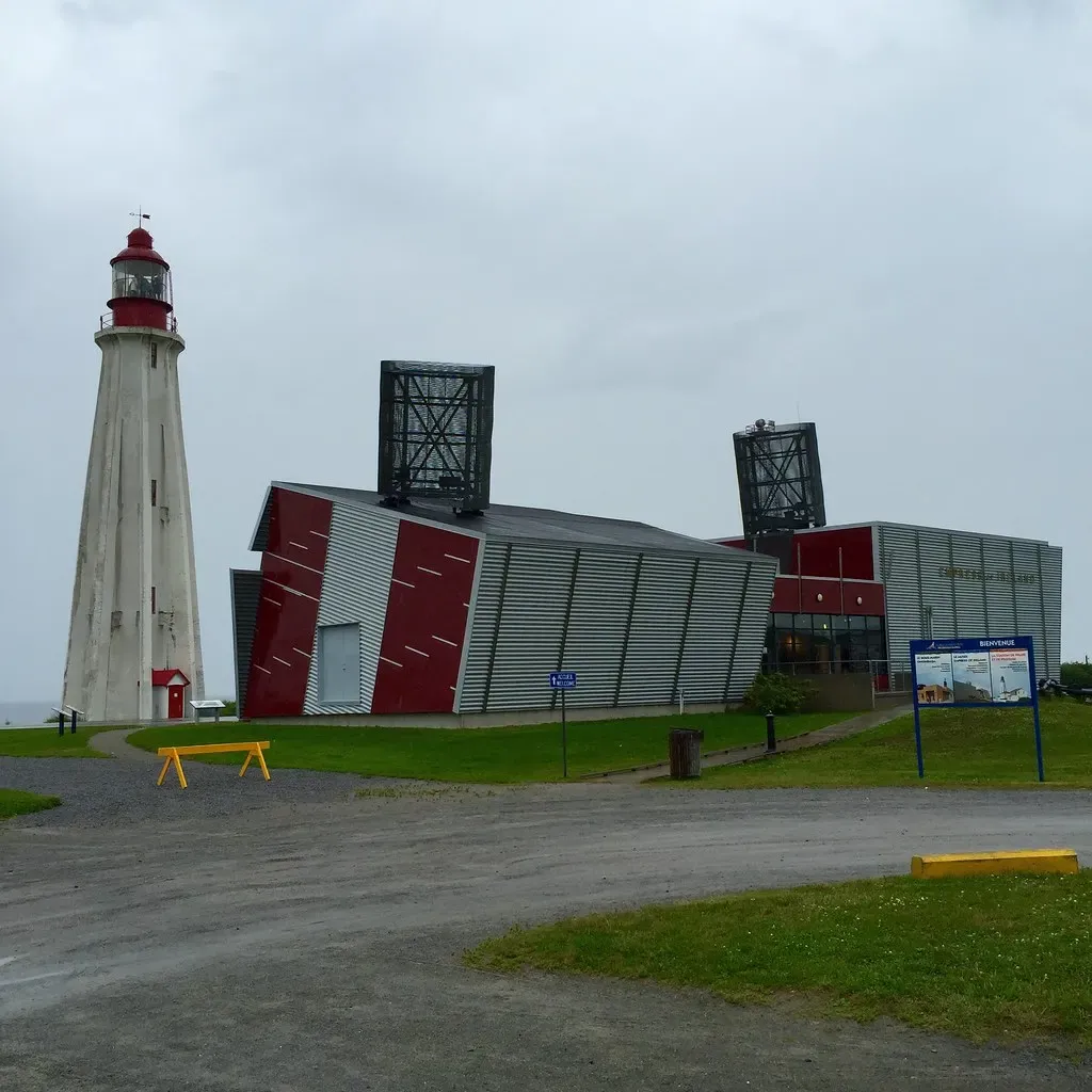 Concrete lighthouse beside a red and grey pavilion building under an overcast sky — Rimouski