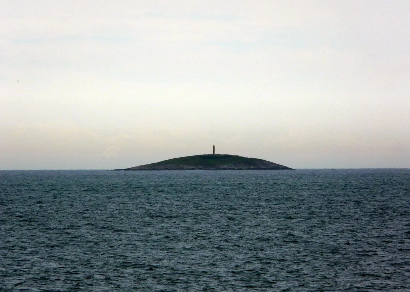 A low, dark island topped by a small lighthouse, seen across grey water under a pale sky — Ilha Escalvada, Guarapari.