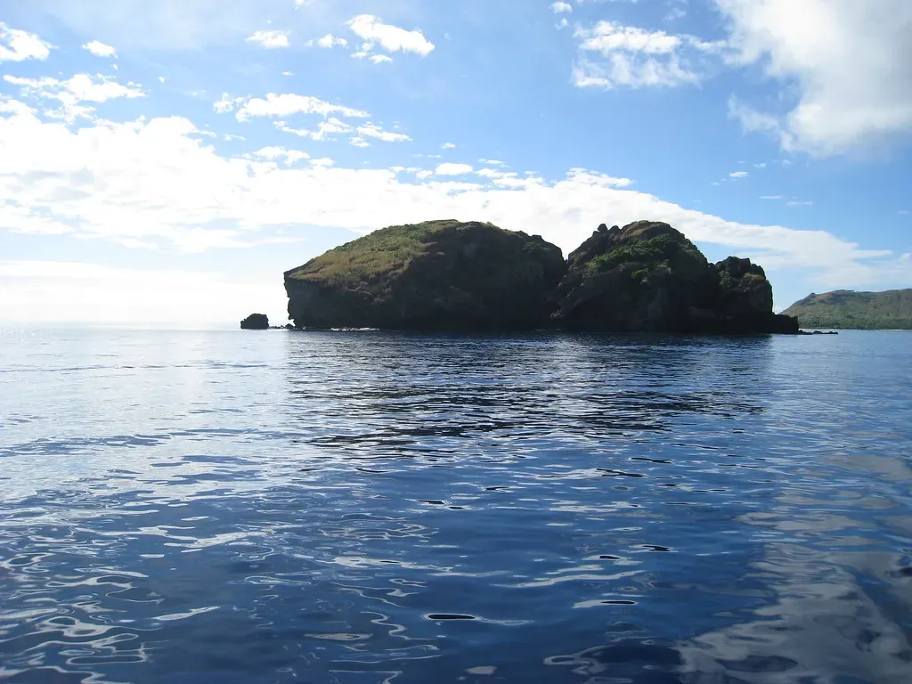 A small rocky islet topped with green scrub sits in calm blue sea under a partly cloudy sky — Yasawa Islands