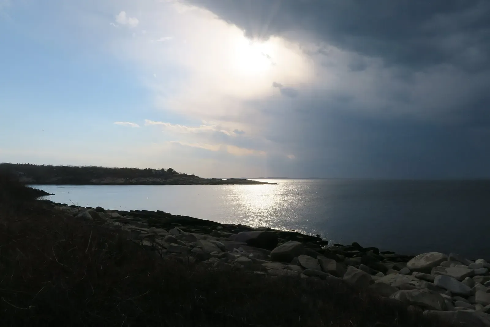 A rocky granite shore curving round a calm bay under a stormy sky with sun breaking through — Folly Cove, Cape Ann & Stellwagen Bank