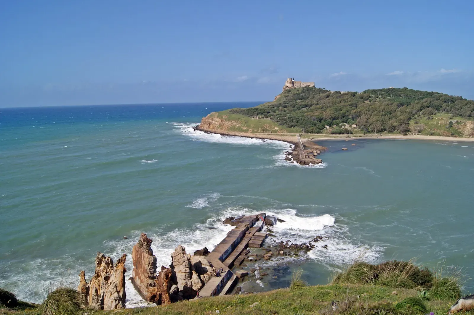 Weathered wreck timbers on foreshore rocks with the headland fort visible across the bay — Cap Tabarka, Tabarka.