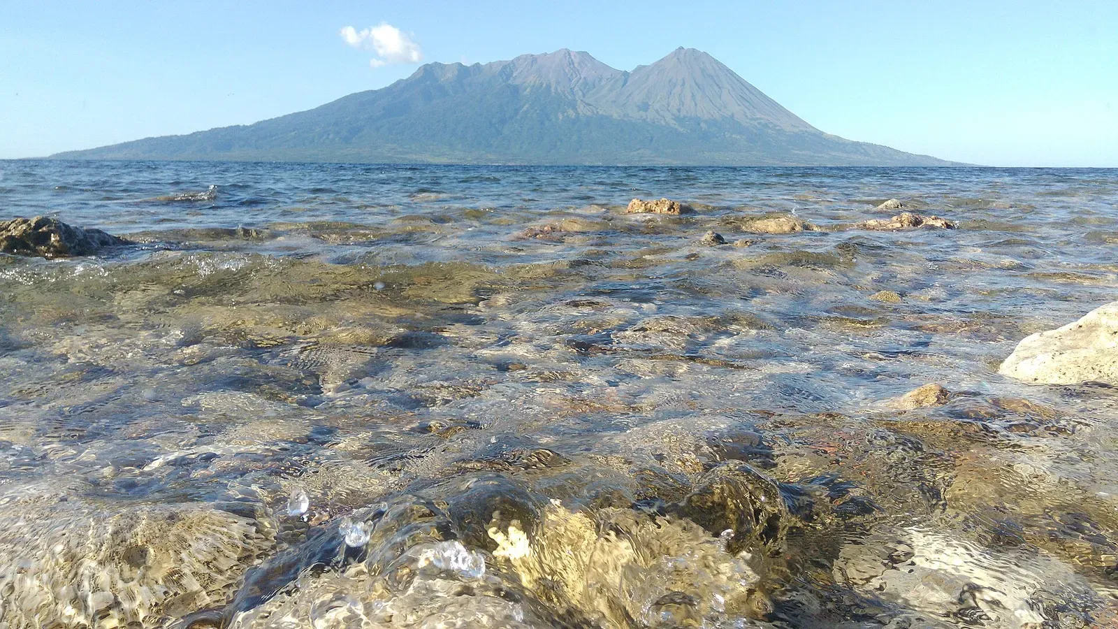 A twin-peaked volcano rising across calm, clear shallow water over a rocky shore under a blue sky — Sangeang & Bima Bay