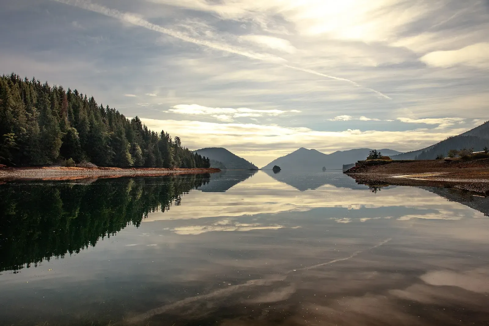 Still water mirrors a forested shore, distant hills and a streaked bright sky — Haida Gwaii
