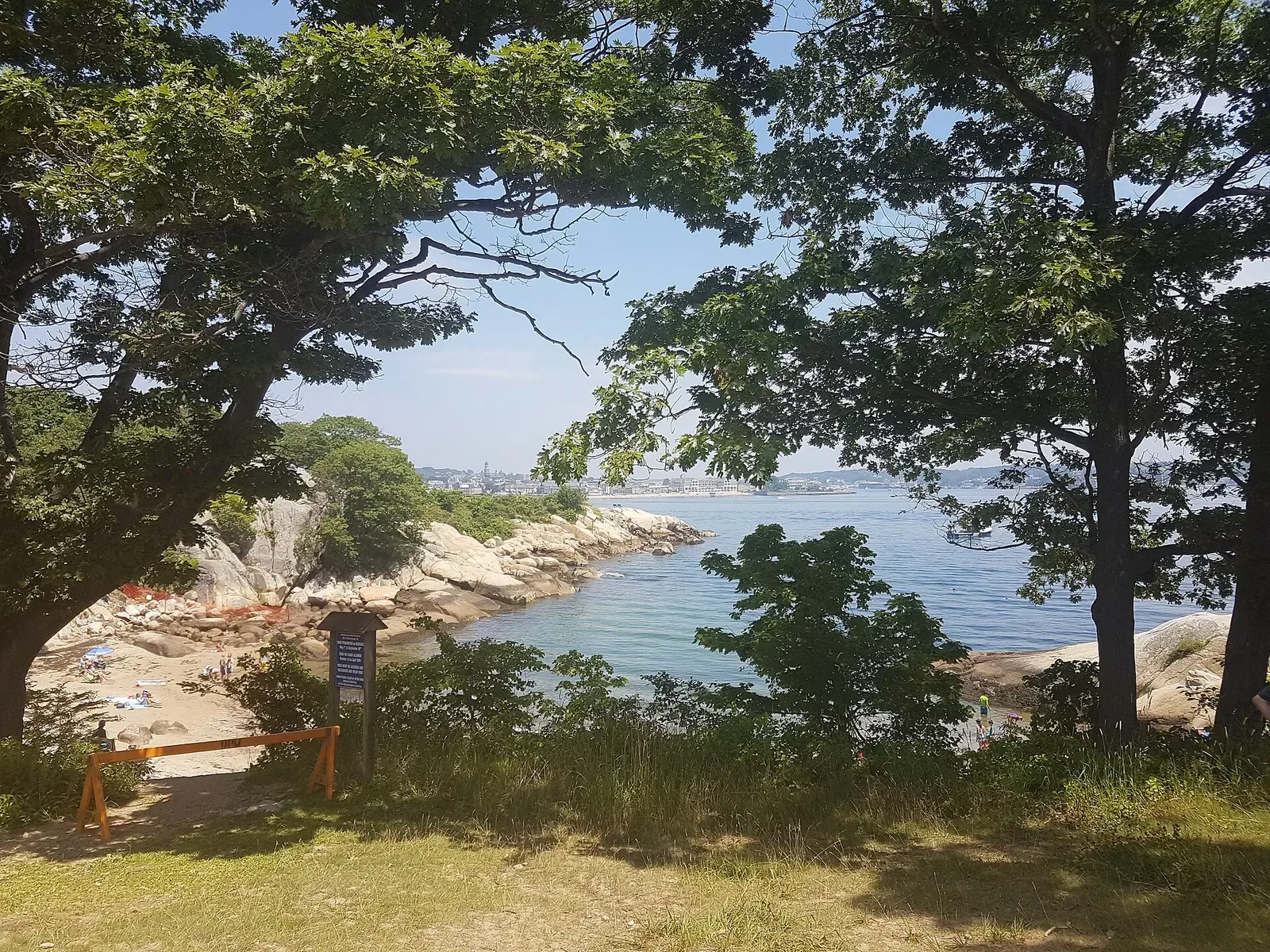 A small sandy cove framed by granite boulders and trees, with Gloucester across the water — Cape Ann & Stellwagen Bank