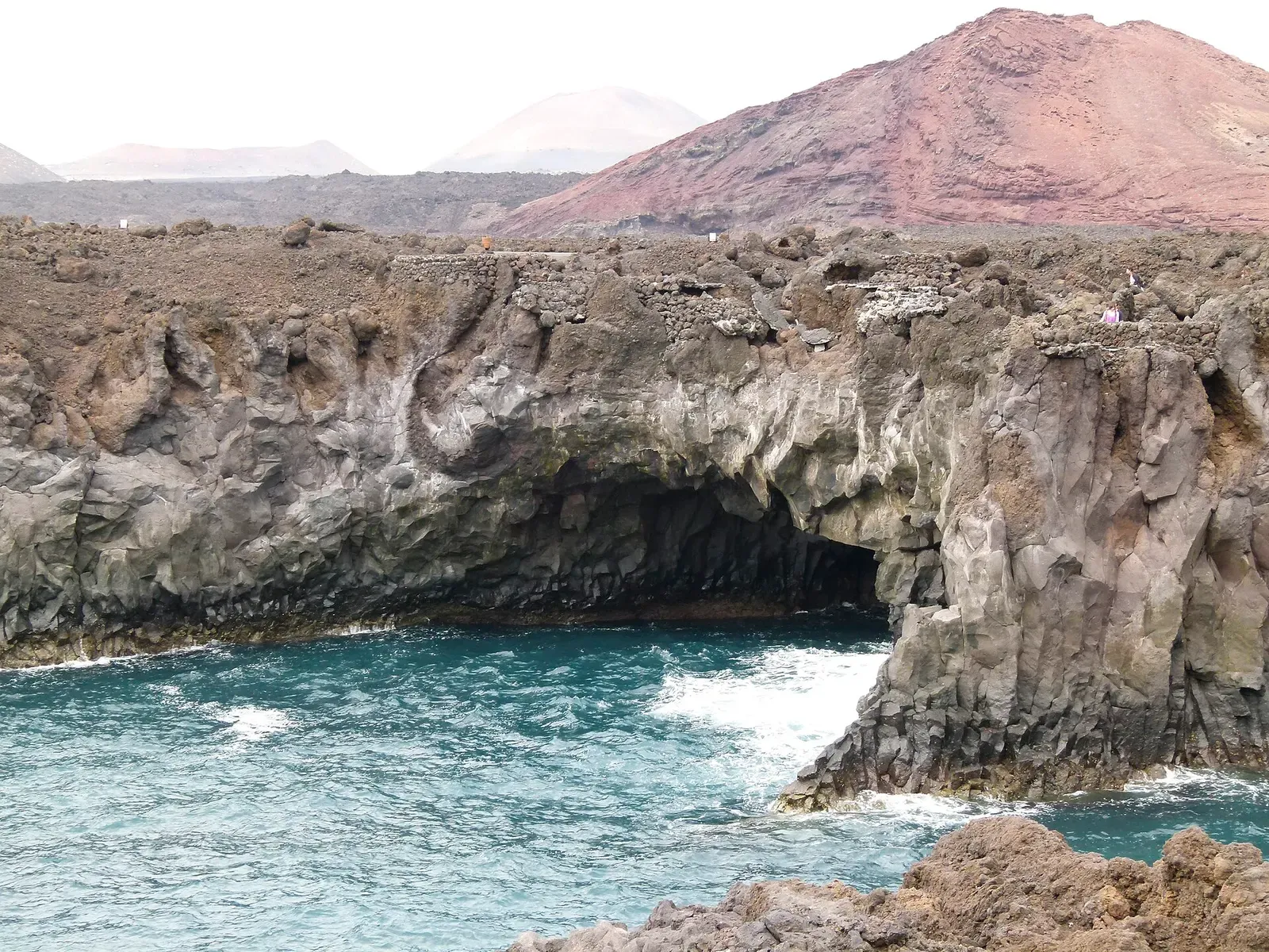 Waves surge into a lava sea arch below the red volcanic cone of Montaña Bermeja at Los Hervideros — Lanzarote