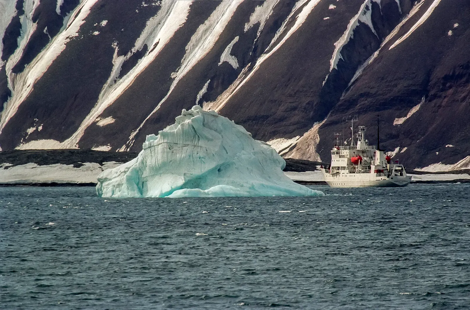 A blue-white iceberg floats near a small expedition vessel below dark mountain slopes in Hornsund — Svalbard.