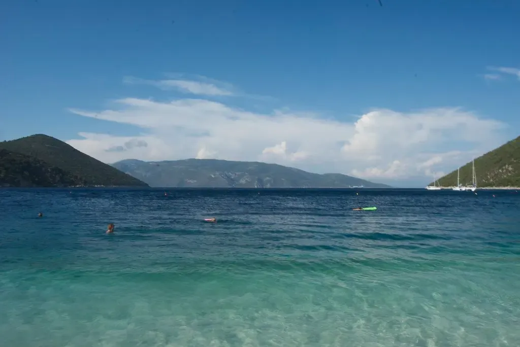 Clear turquoise shallows in front of green hills across the water, with two moored sailing boats on the right — Kefalonia & Ithaca