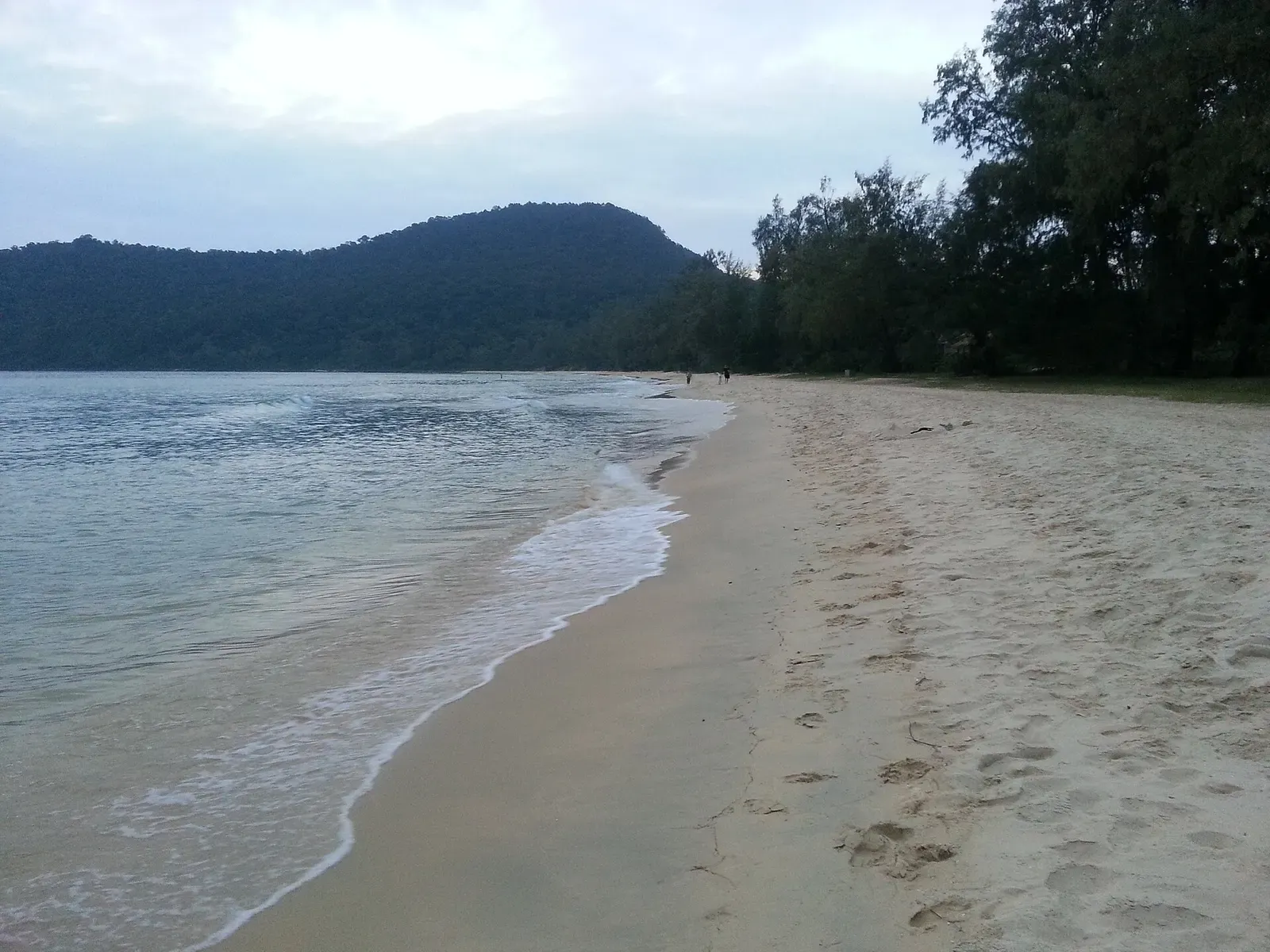 Empty sand beach on Koh Rong Samloem with footprints in the sand and a forested hill behind.