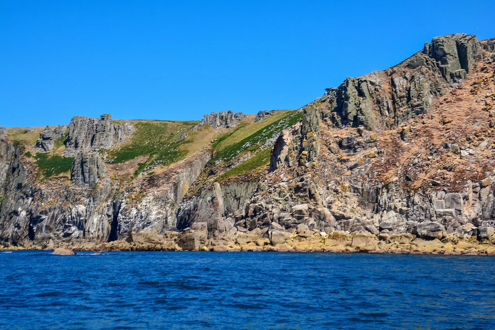 Weathered granite cliff face with boulders at the waterline and a slope of grass above, seen from a boat — Lundy Island