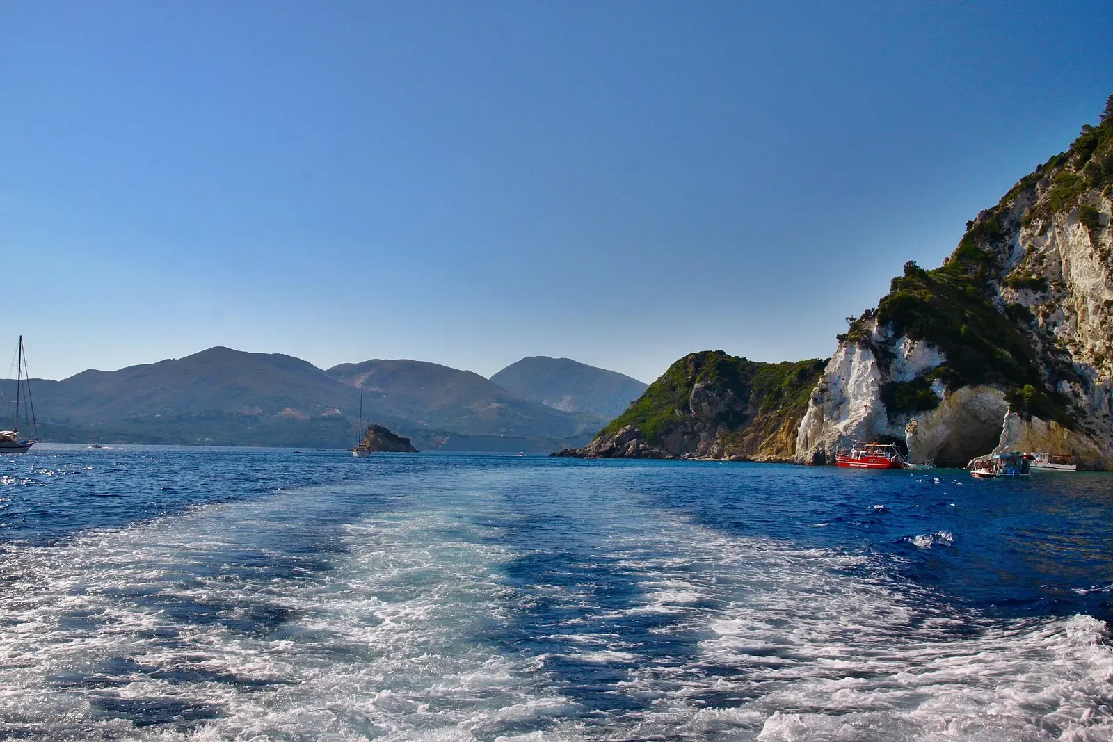 View from a boat's wake toward Marathonisi island under a clear sky.