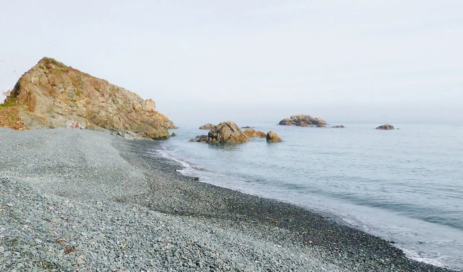 A grey shingle beach running to a rocky point, with a few rocks offshore in a calm, misty sea — Cornwall