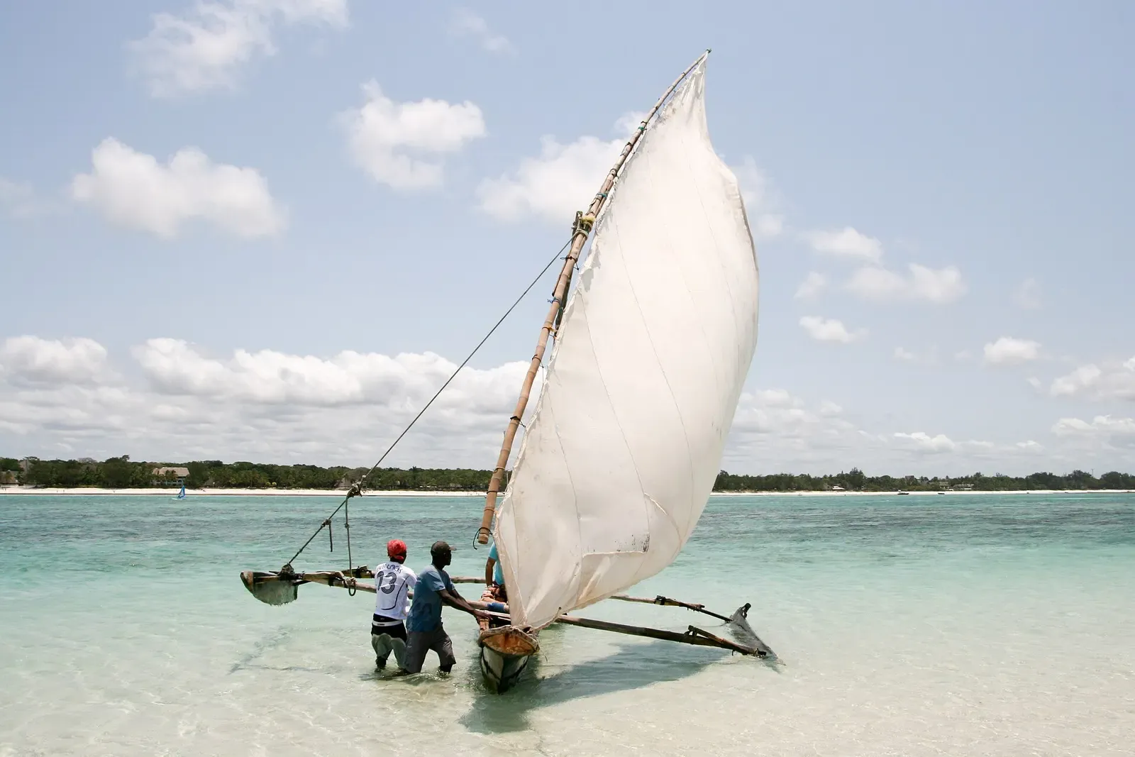 Two men push a wooden outrigger canoe with a large white sail through shallow turquoise water, with a low wooded shore behind — Diani Beach.