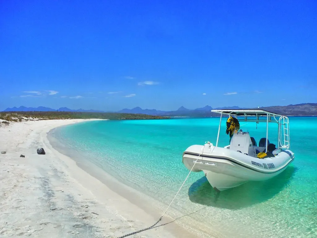 A white inflatable dive boat moored off a white sand beach in turquoise shallows, desert mountains behind — Loreto Bay National Park
