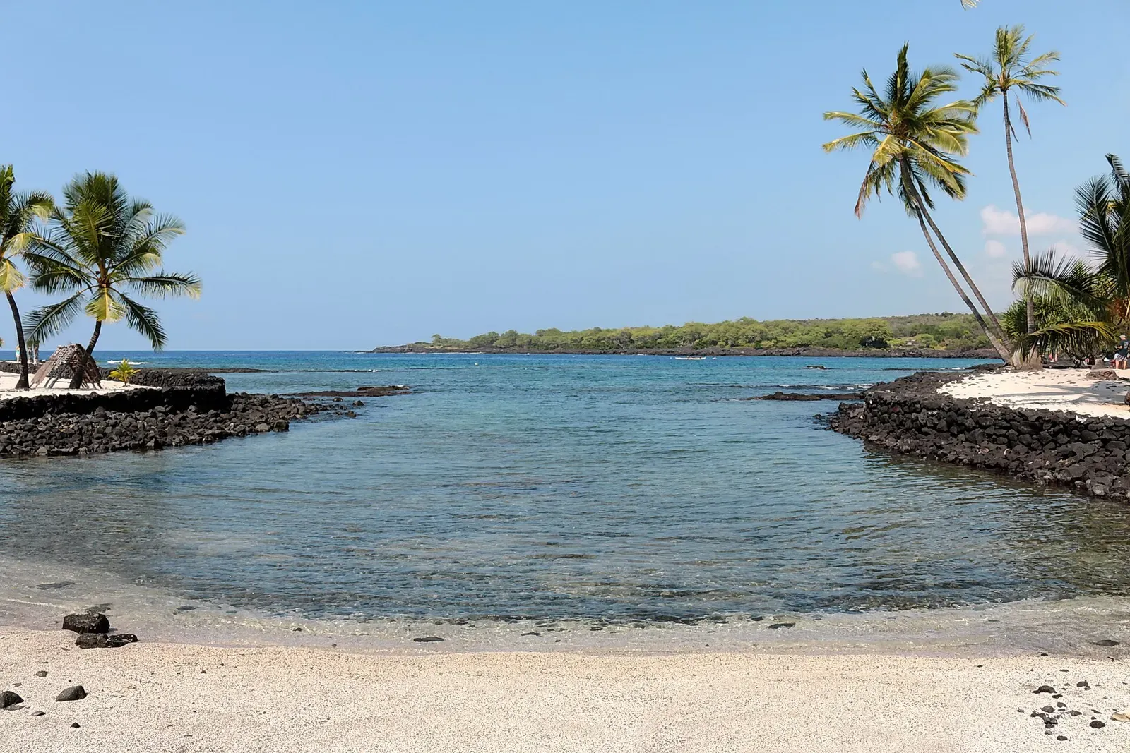 A calm cove of clear blue water meets a narrow sand and lava shoreline at Honaunau Bay — Kona.