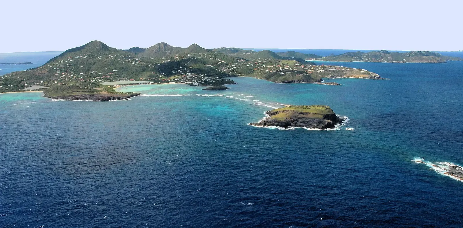 Aerial view of a green, hilly island with turquoise bays and a small rocky islet in deep blue water in front — Saint Barthélemy.