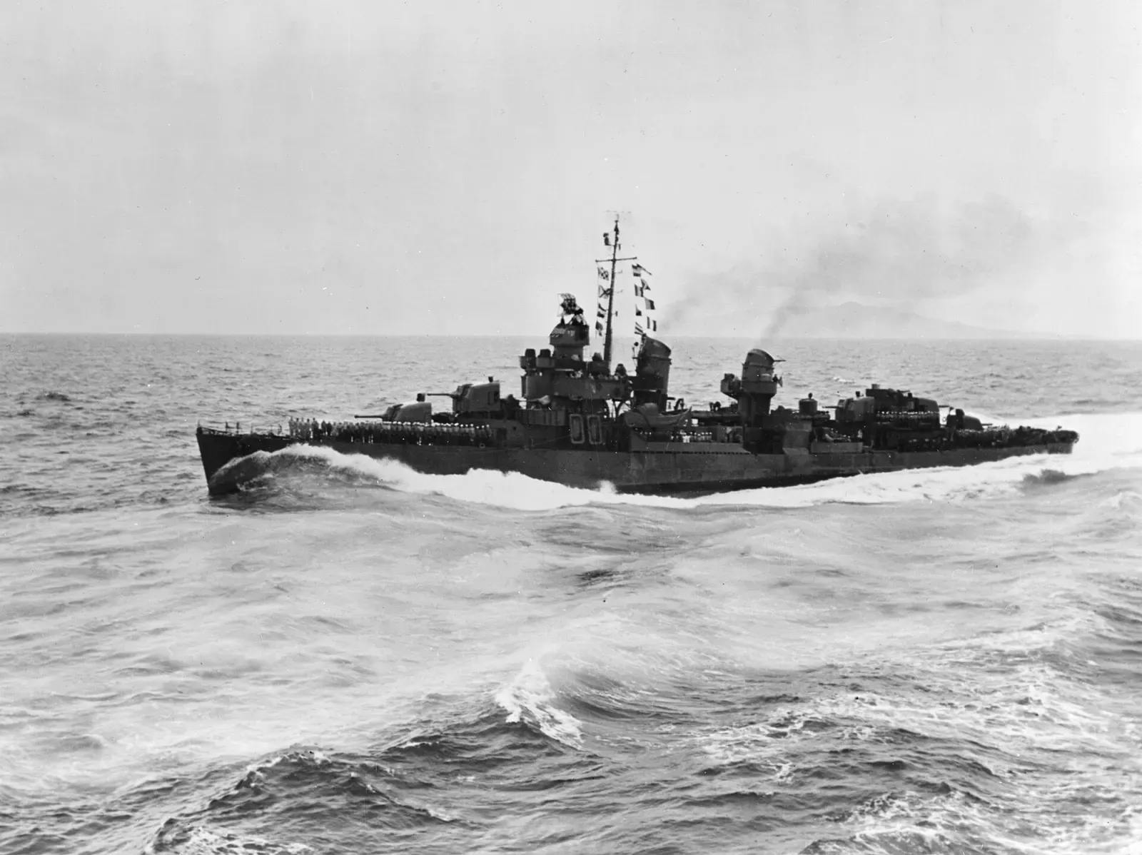 Black-and-white photograph of a US Navy destroyer with crew lining the rails, throwing spray in a heavy swell with land on the horizon — Iron Bottom Sound