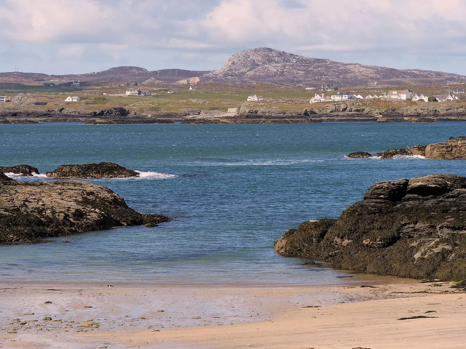 Blue water of a bay framed by dark rocks, with a rocky hill and houses on the far side — Porth Castell, Anglesey