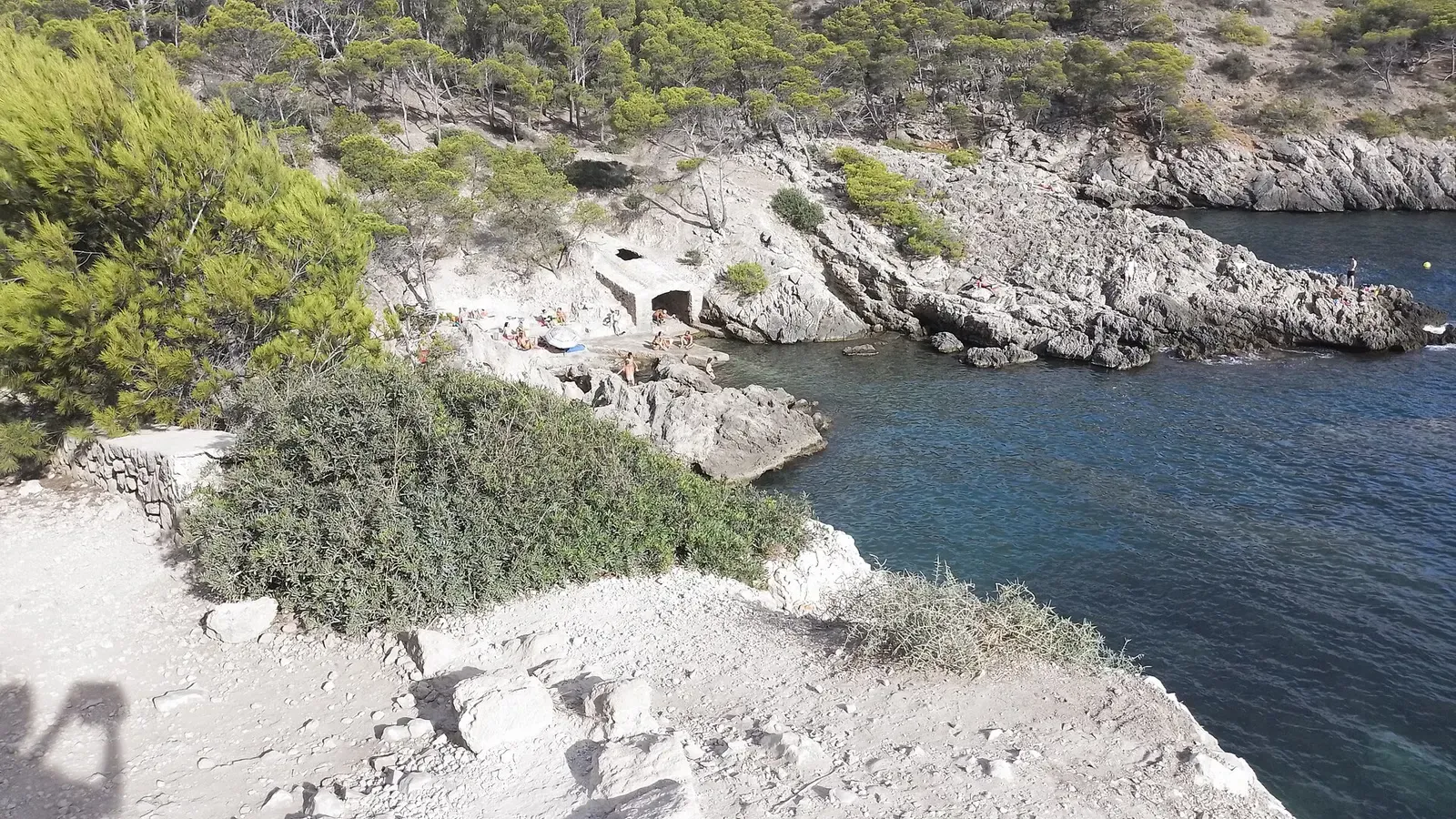 View across Caló des Monjo towards the sea, with a dark cave opening partway up the far cliff and a small stony beach