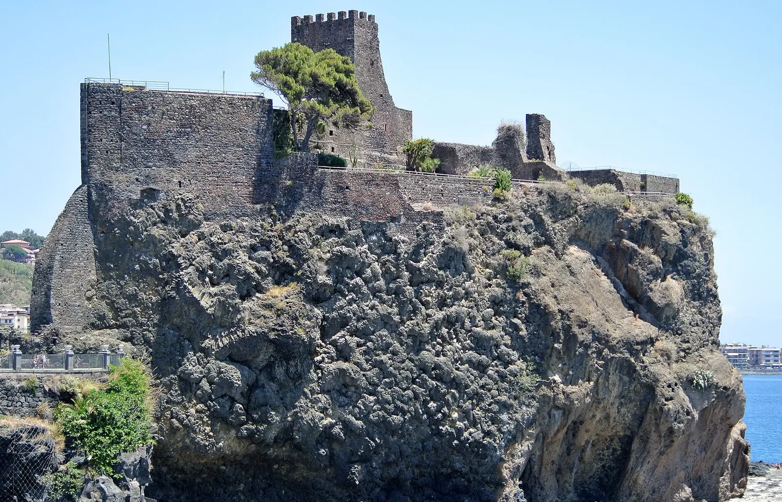 A medieval castle with a crenellated tower sits atop a dark volcanic-rock cliff above the sea.