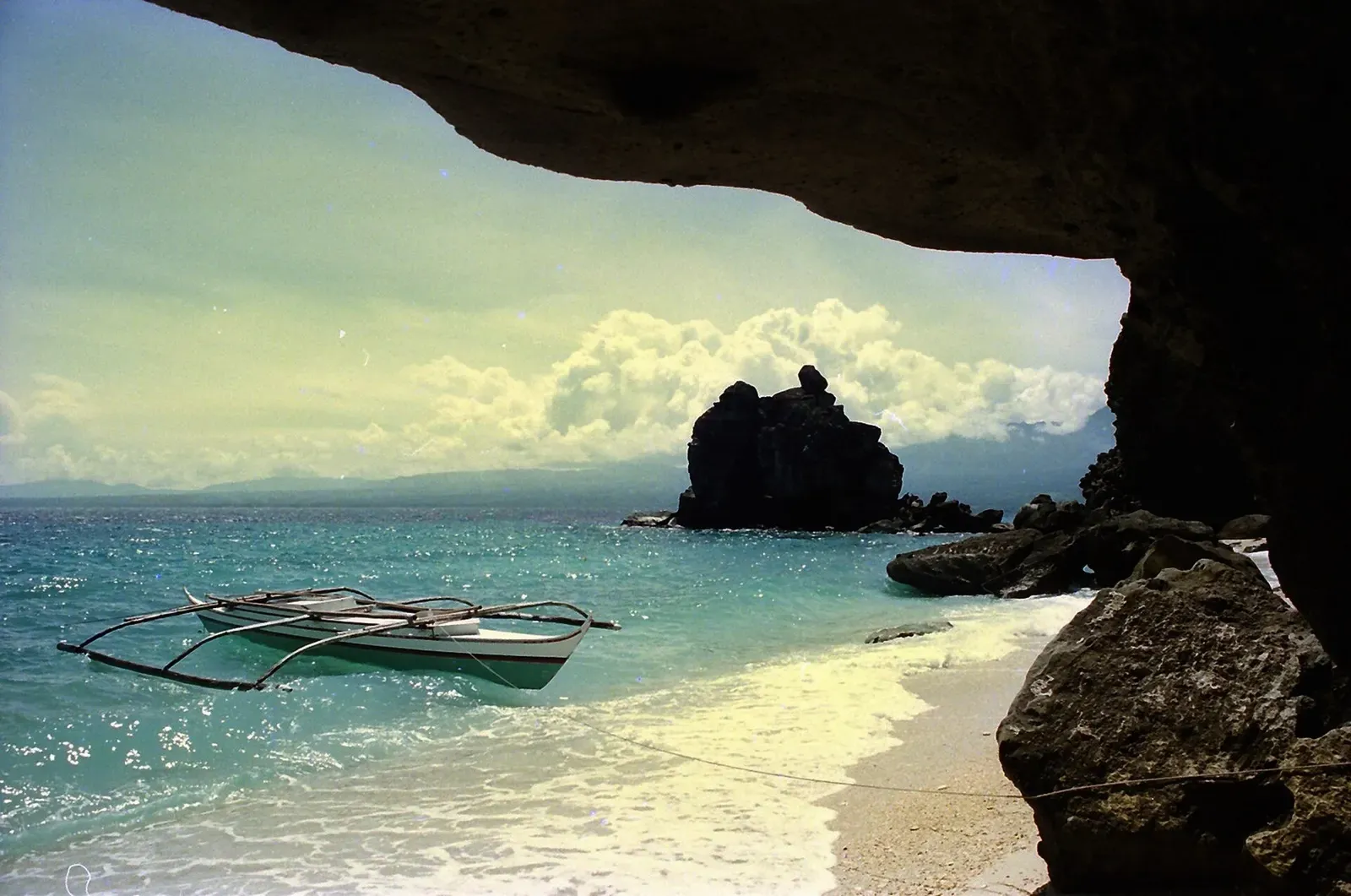An outrigger boat moored off a white beach on Apo Island, seen from under a rock overhang with a sea stack beyond — Katipanan Reef.