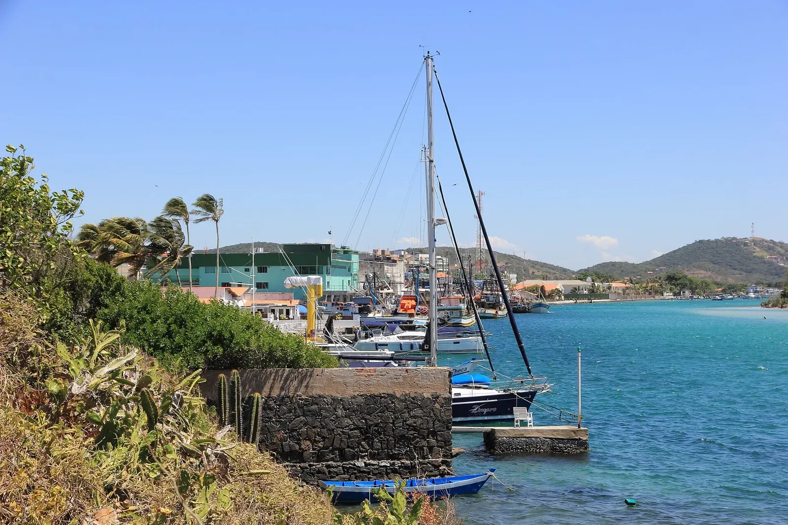 Sailing yachts and small boats moored along a stone quay in a calm blue channel with green hills behind — Cabo Frio