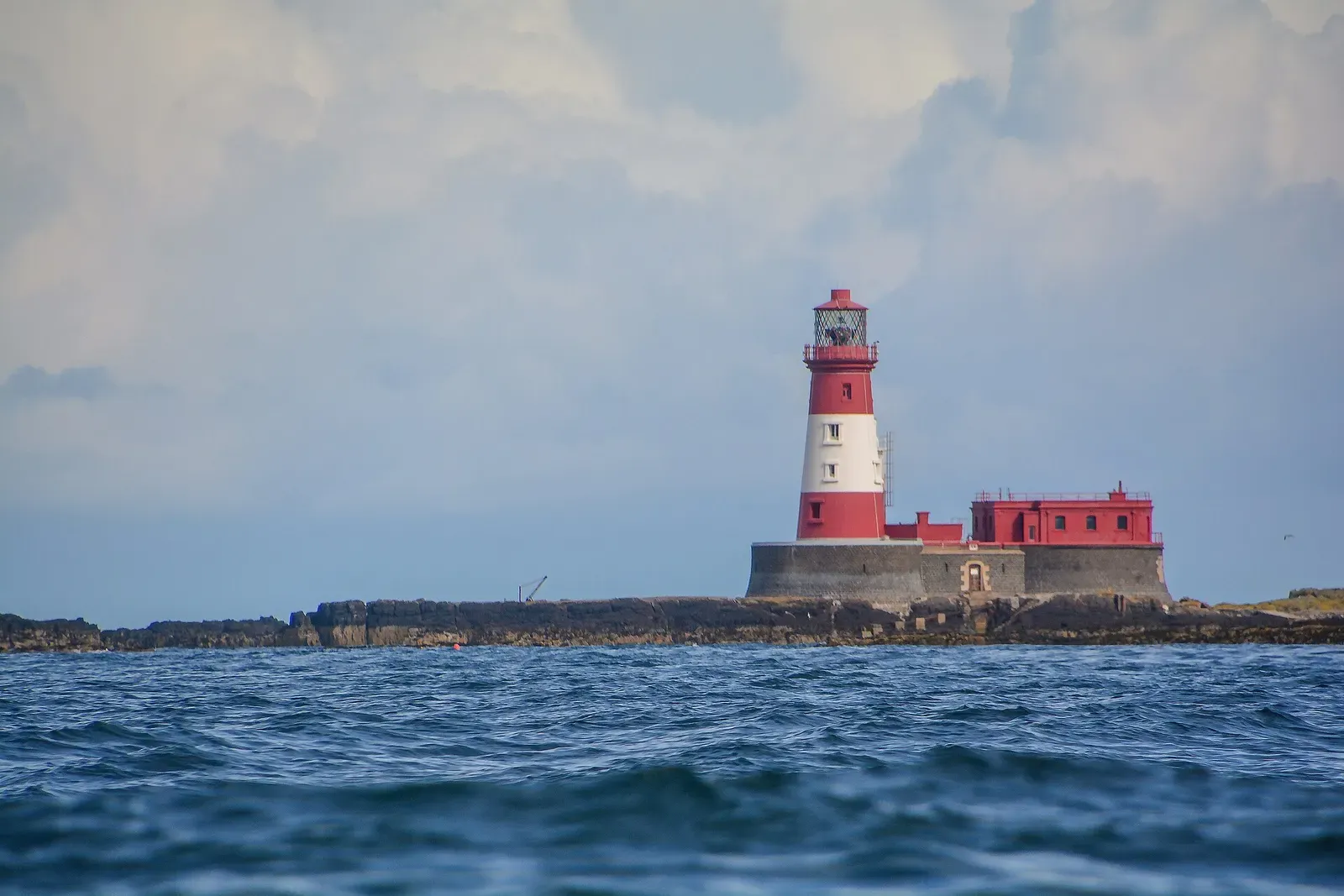 Longstone Lighthouse under a dramatic cloudy sky, seen across the exposed rocky reef of the Farnes — Farne Islands.