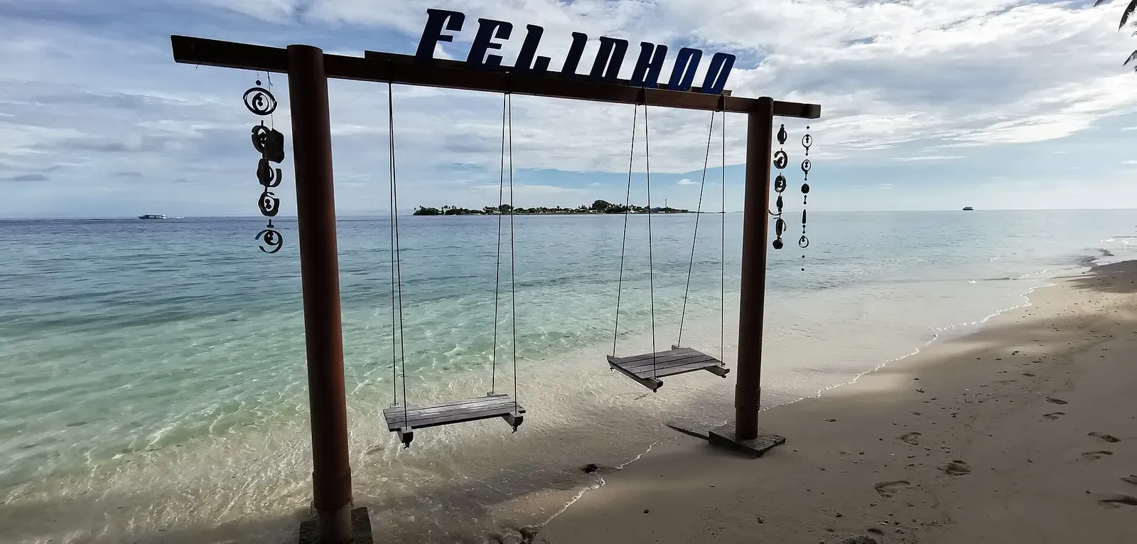 A wooden beach swing set on the sand with a Felidhoo sign, facing turquoise shallows