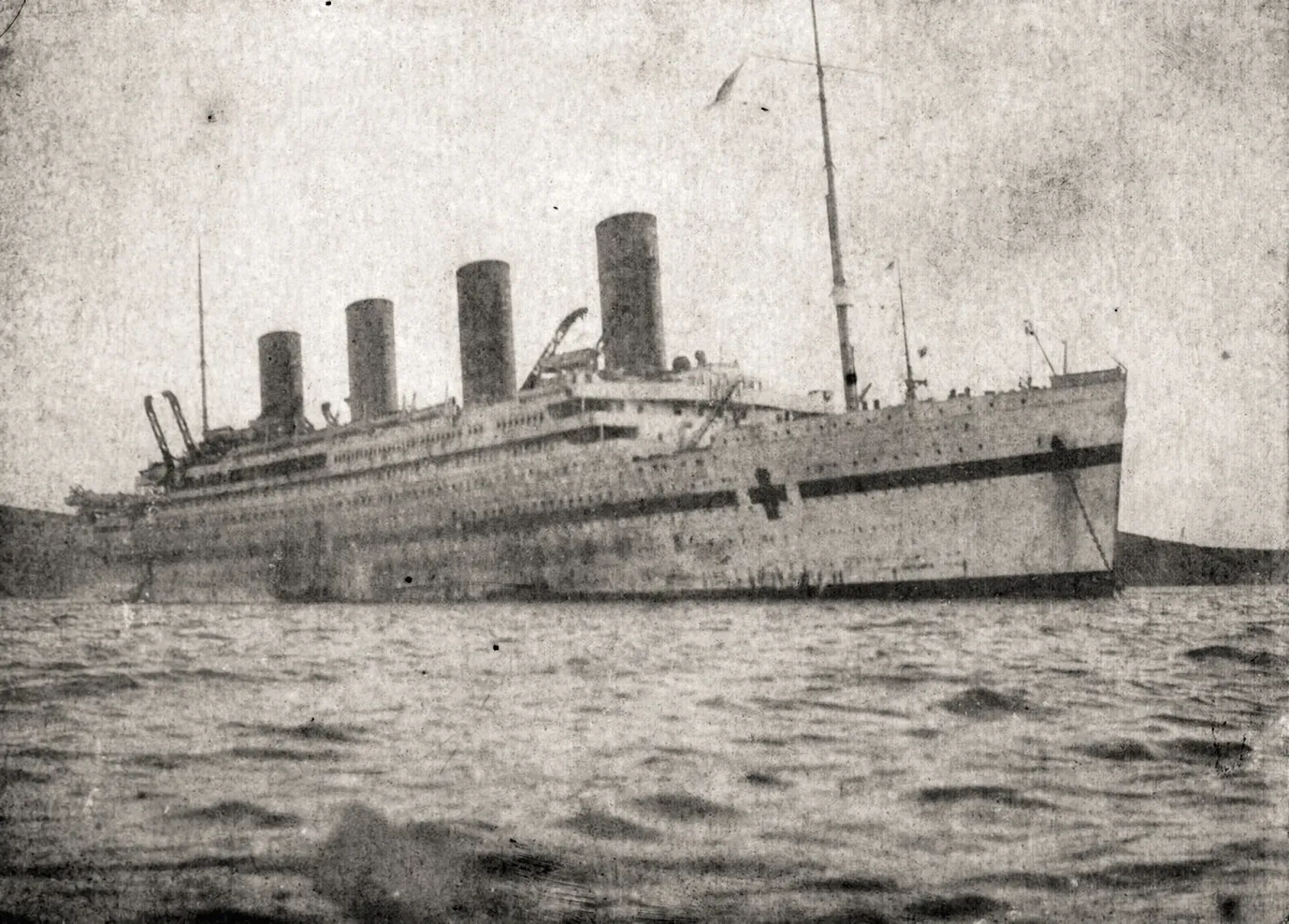 Weathered black-and-white photograph of HMHS Britannic at anchor, her four funnels and hull with hospital-ship red cross markings reflected in calm water.