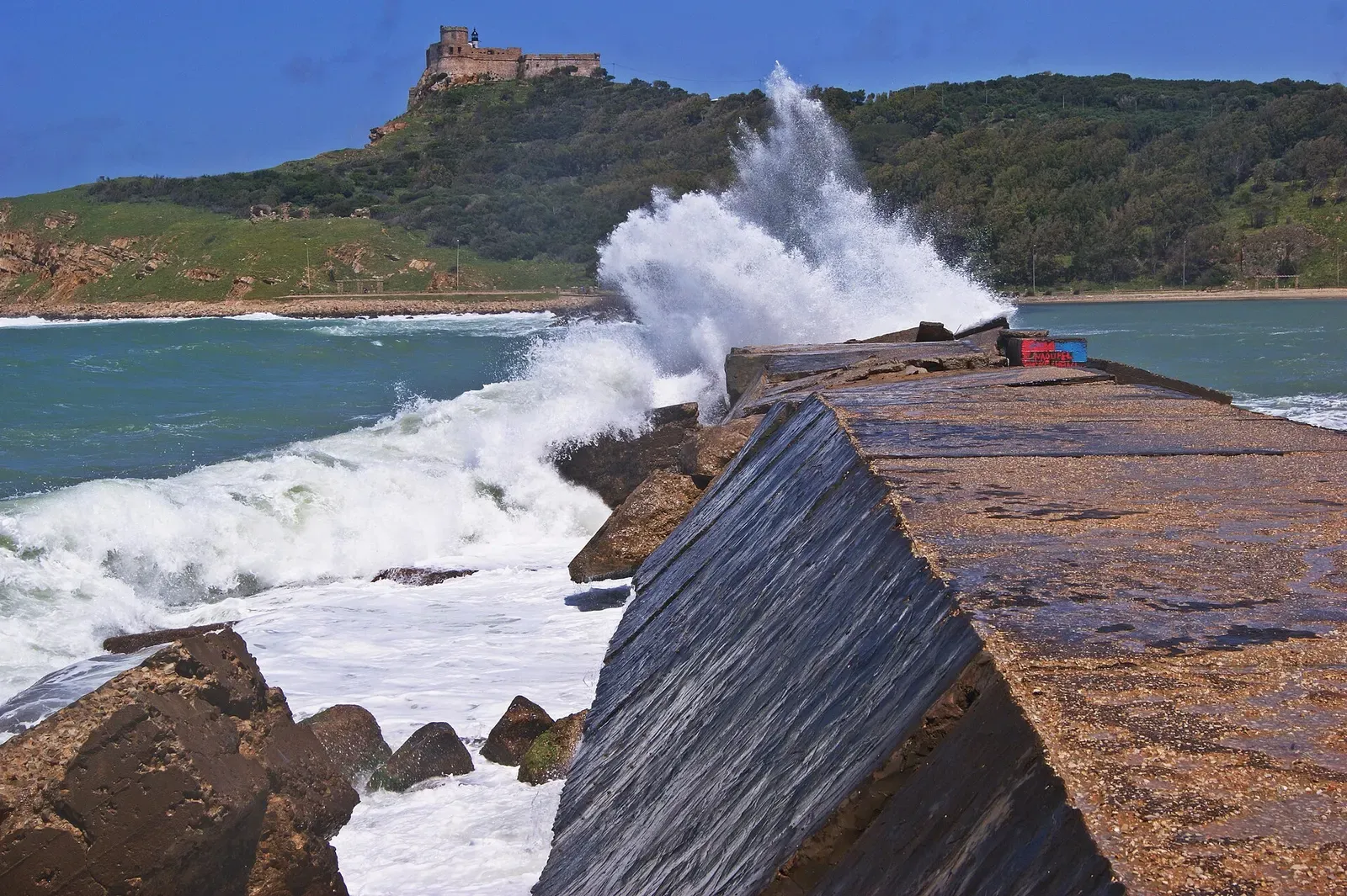 Waves breaking over Tabarka's harbour breakwater with the headland fort behind — Cap Tabarka.
