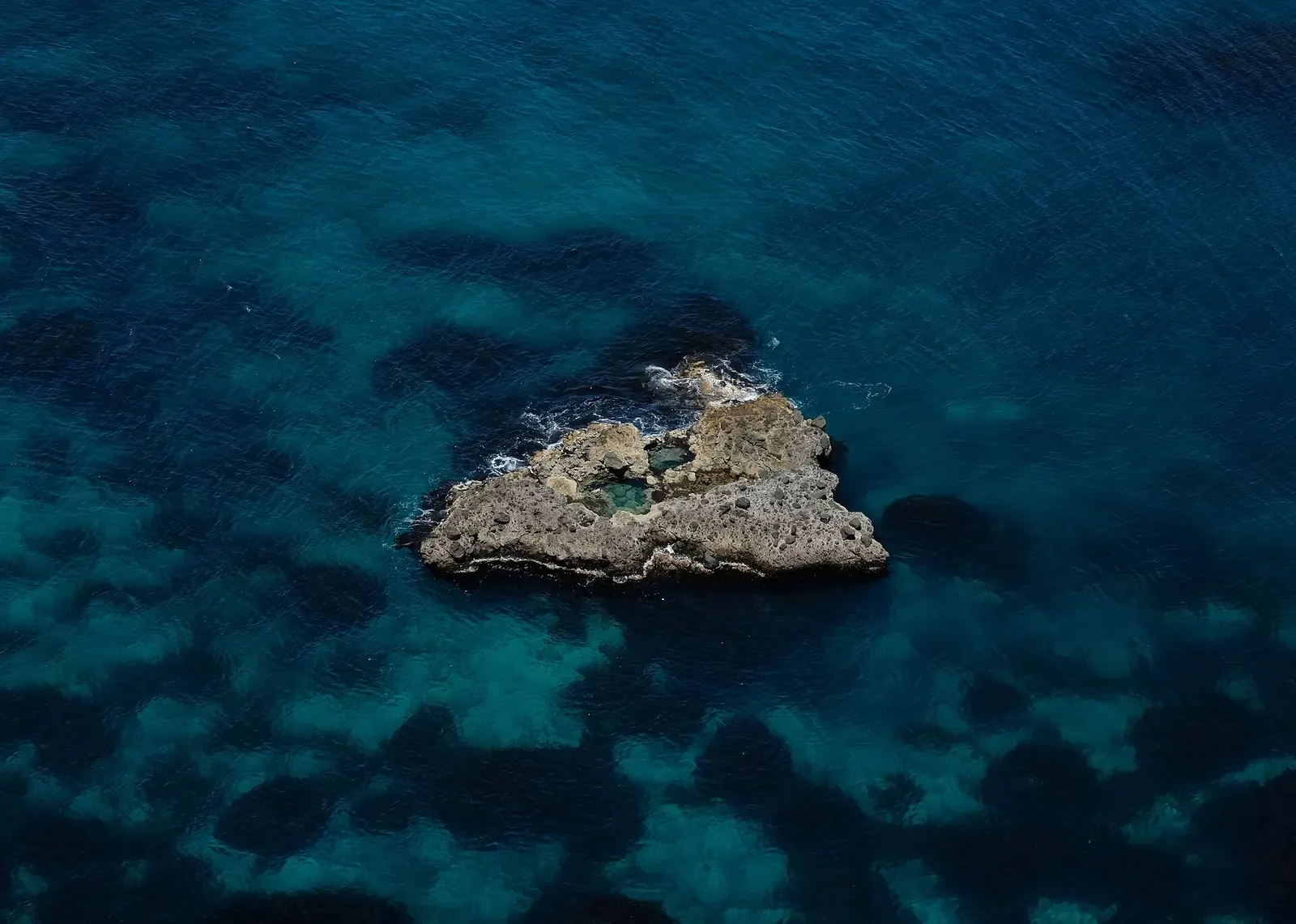Aerial view of a small flat rocky islet surrounded by deep blue water with dark rock patches below — Shakotan Peninsula
