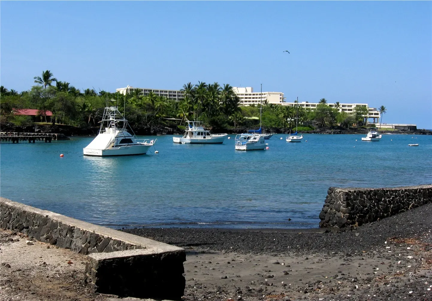 Charter and sport-fishing boats moored in Keauhou Bay below the resort on the point, with a lava-walled boat ramp in the foreground — Kona