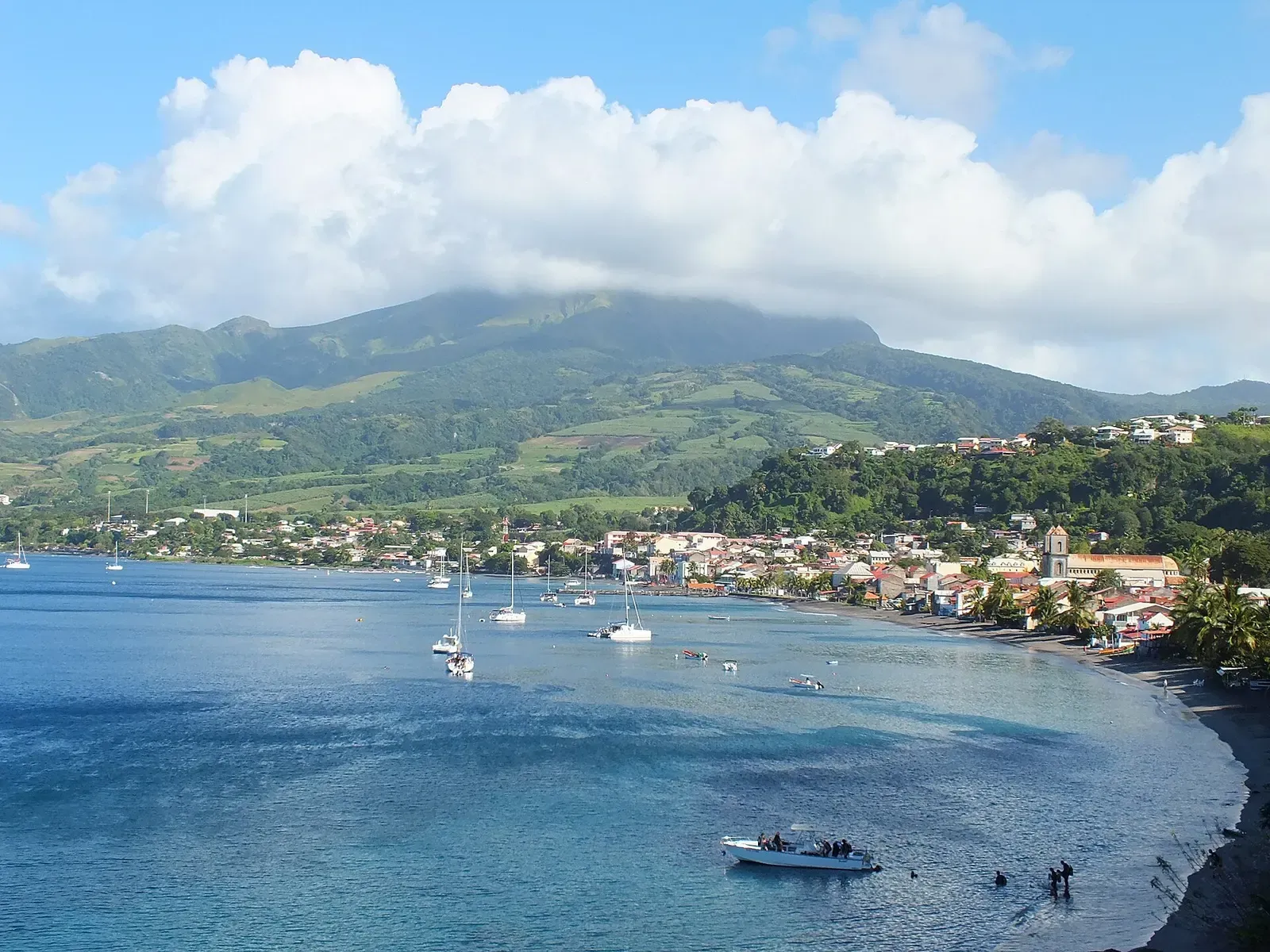 A blue bay with moored sailboats and a shoreline town below a green, cloud-capped mountain — Martinique