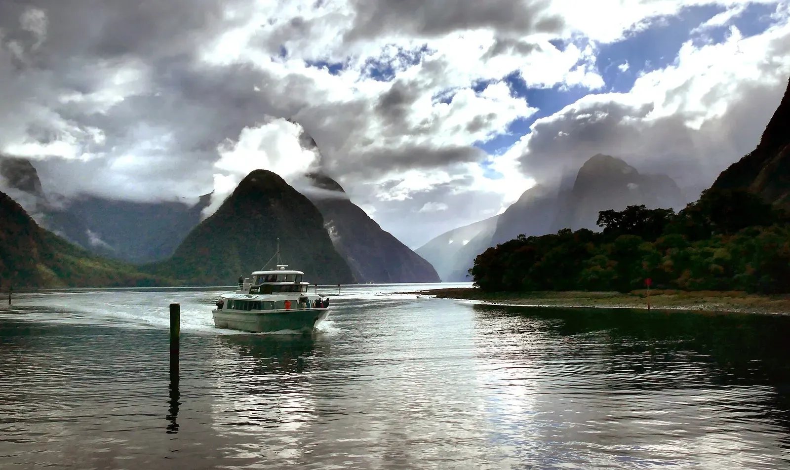 Cloud-wreathed peaks above Milford Sound with a small cruise boat entering the fiord — Fiordland