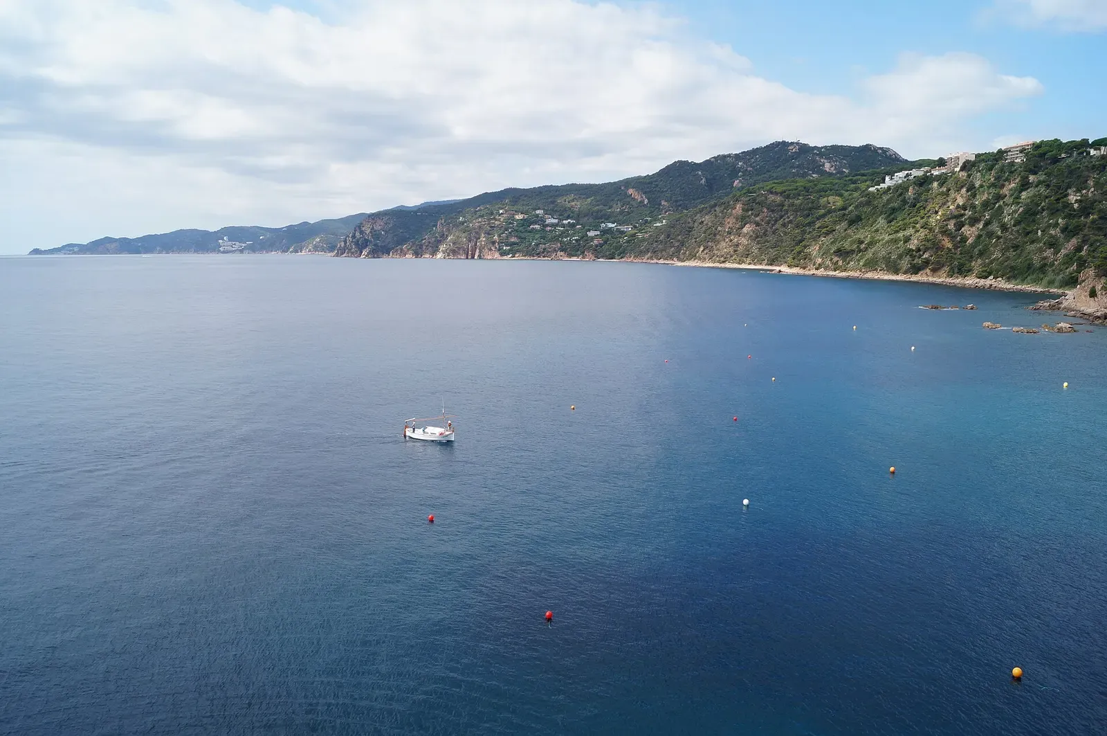 A small boat moored in the bay below the Mirador de les Modistes, wooded hills on the far shore
