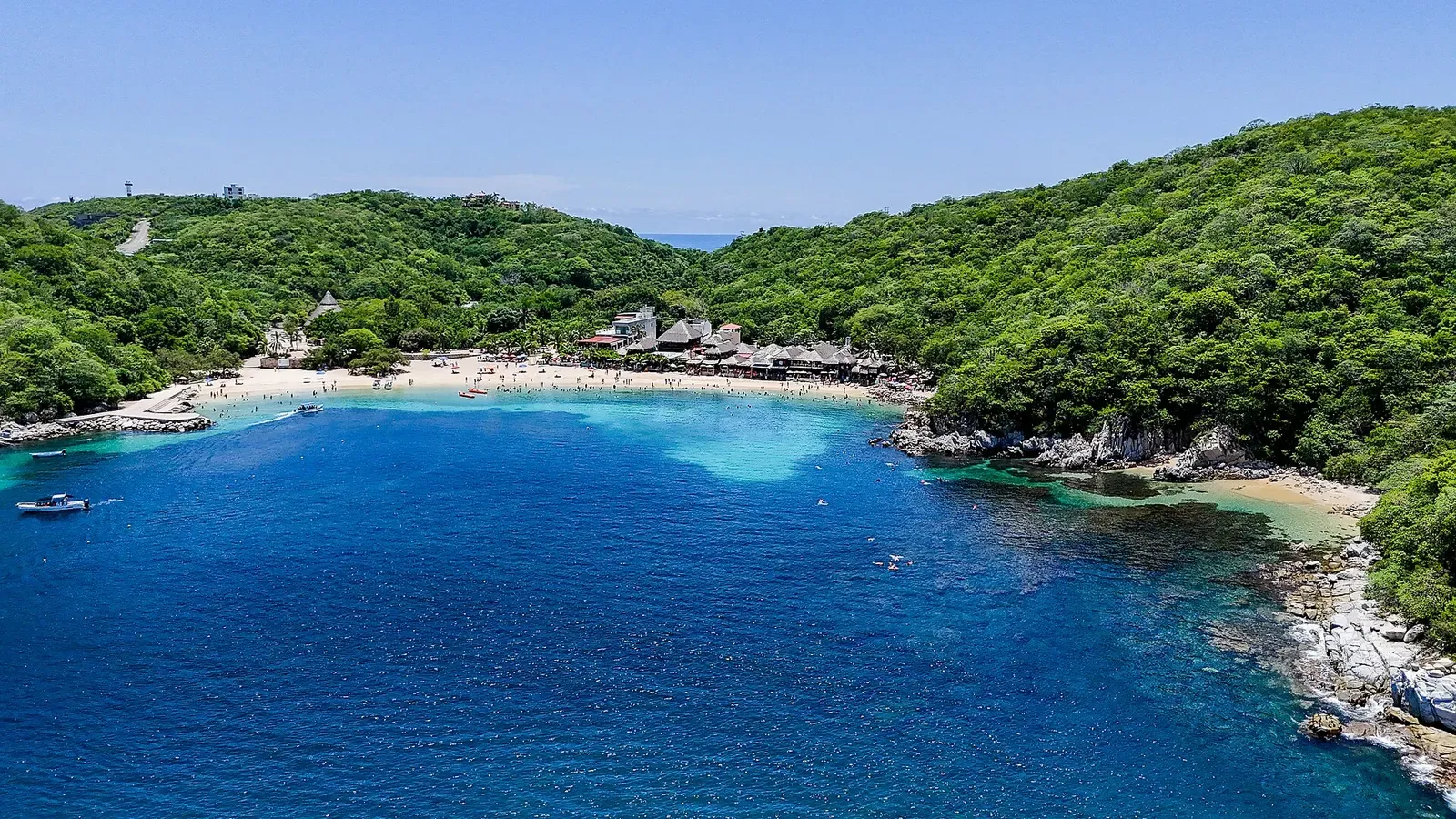 Aerial view of a small cove with a pale sandy shallow, palapa restaurants and forested hills — Huatulco