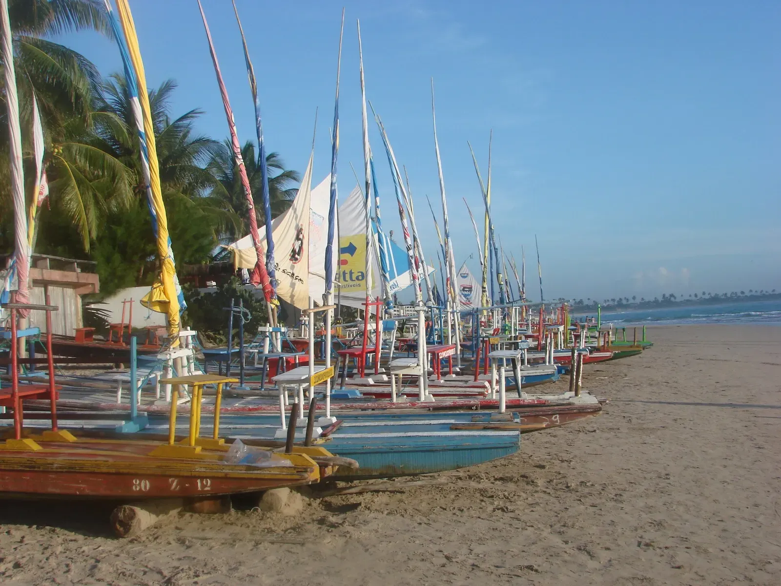 A row of colourful jangada sailboats with tall masts drawn up on a sandy beach — Porto de Galinhas