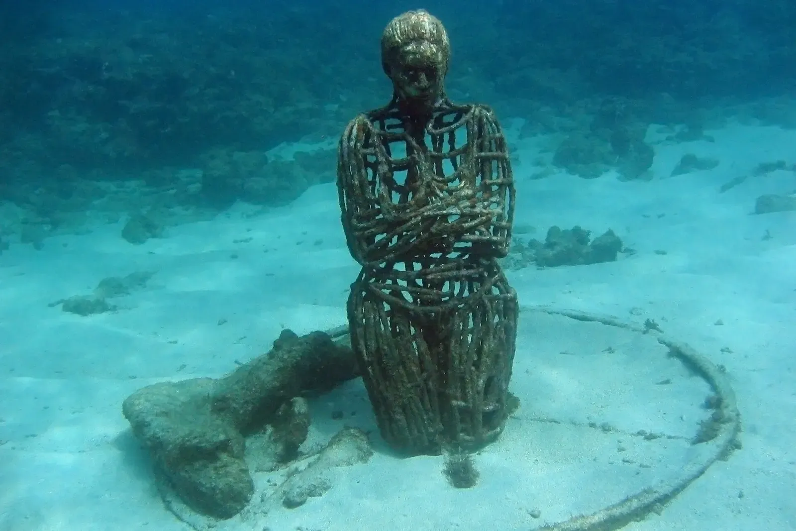 A kneeling cast figure made of wire mesh, chained at the wrists, resting on the sandy bottom — Underwater Sculpture Park, Grenada