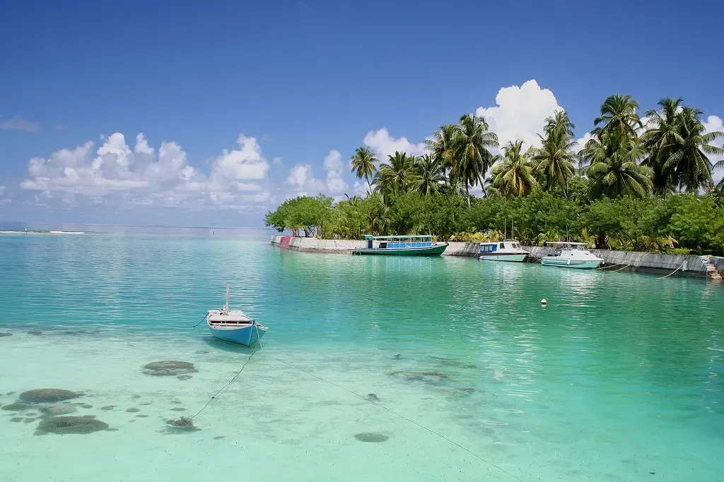 Small boats moored off a palm-lined shore with a jetty and huts in Addu Atoll