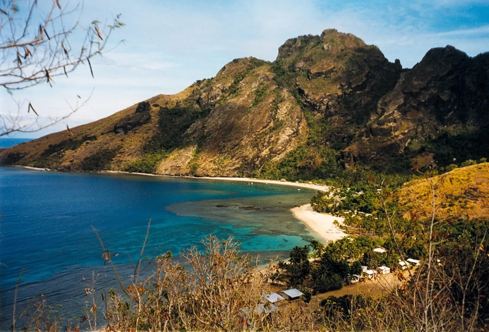A steep rocky peak above a curving bay with a white sand beach, shallow turquoise reef and dry grass in front — Yasawa Islands