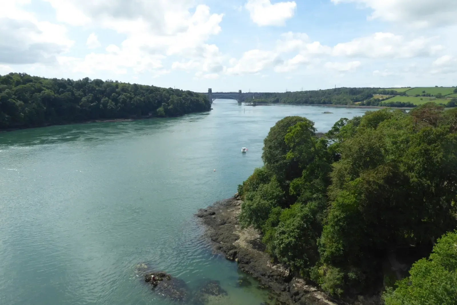 A wooded headland and the turquoise water of a strait, with a distant bridge on the horizon — Menai Suspension Bridge, Anglesey