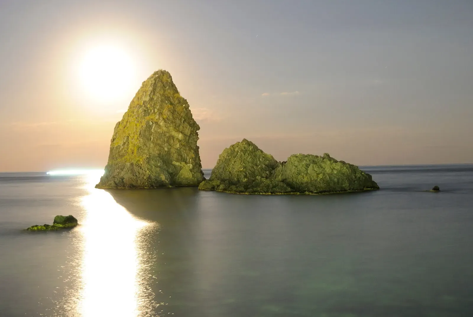 Three moss-covered rock stacks stand together in the sea off the coast in warm evening light.