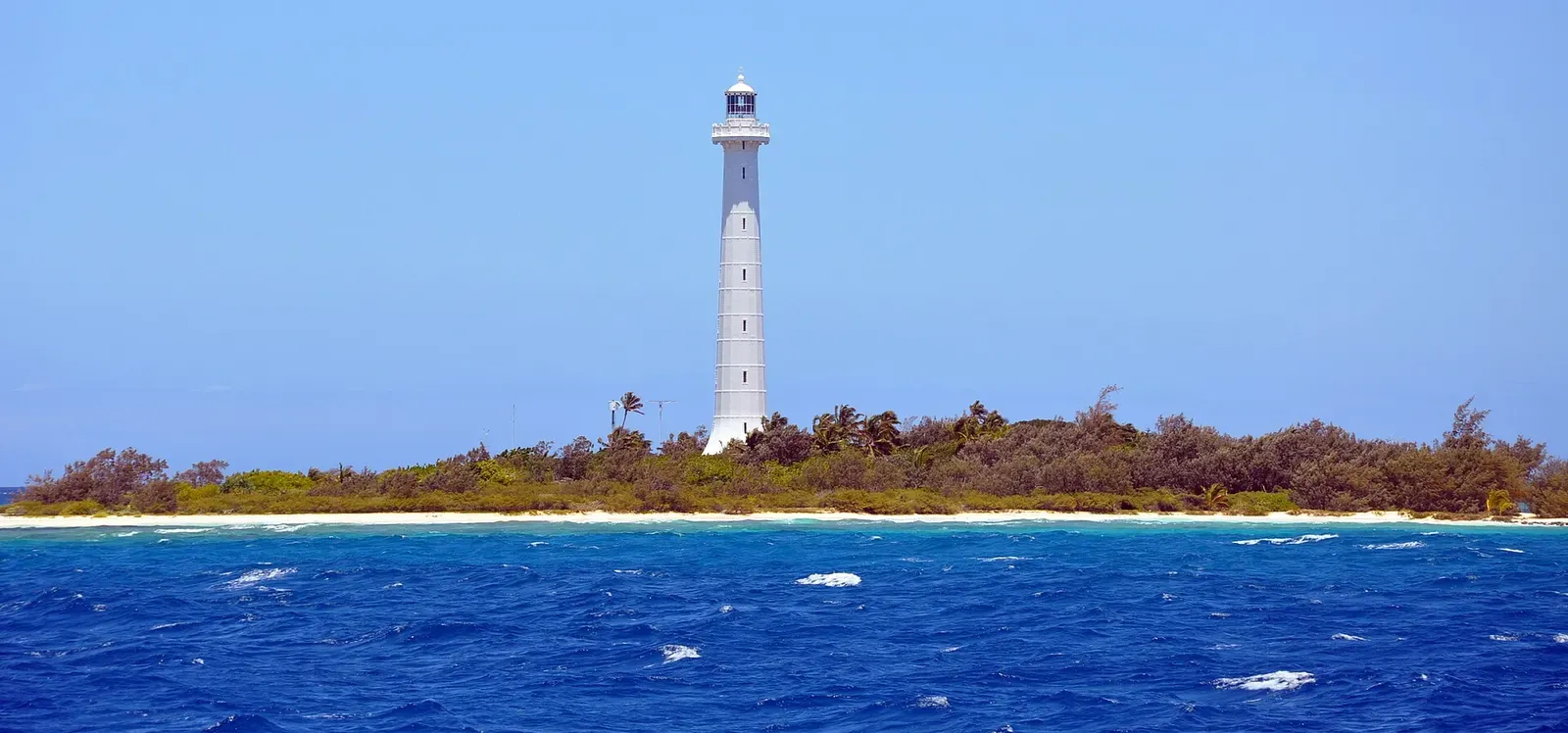 The white Amédée lighthouse rising above the islet's reef, seen from the water — New Caledonia