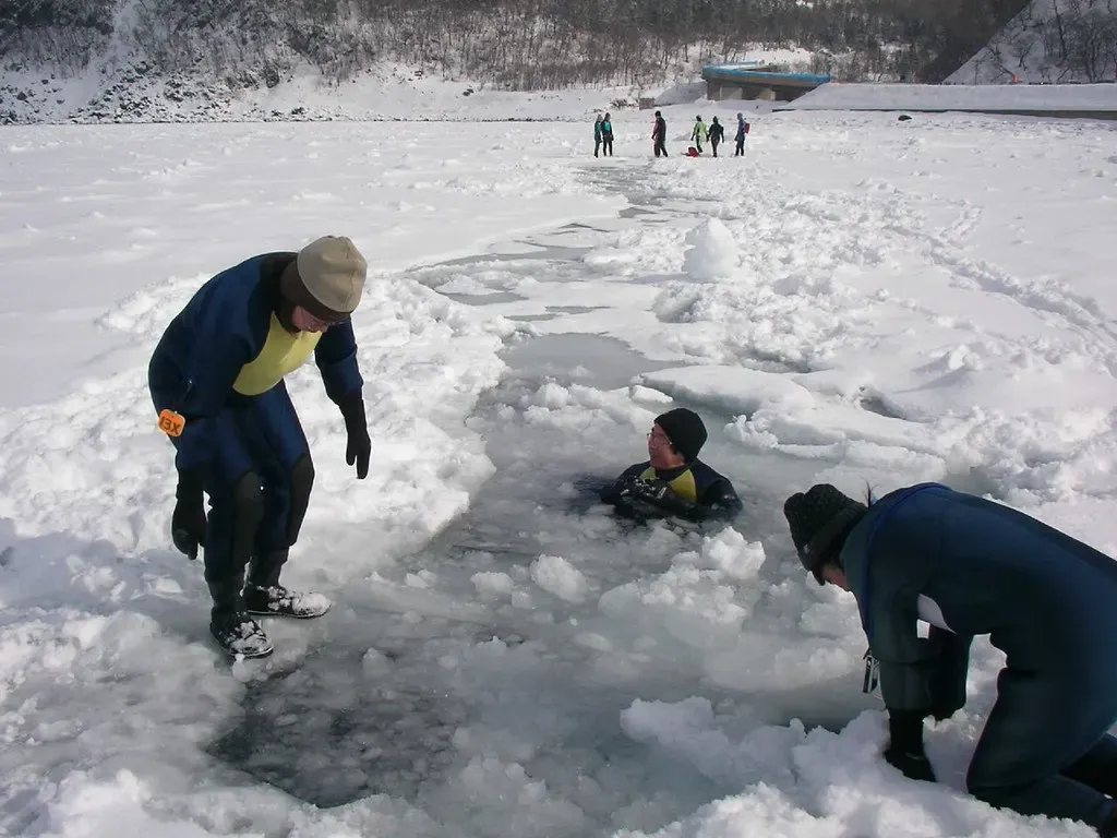 A drysuited person floats in a pool of open water cut into the sea ice while two others look on from the edge — Shiretoko Drift Ice