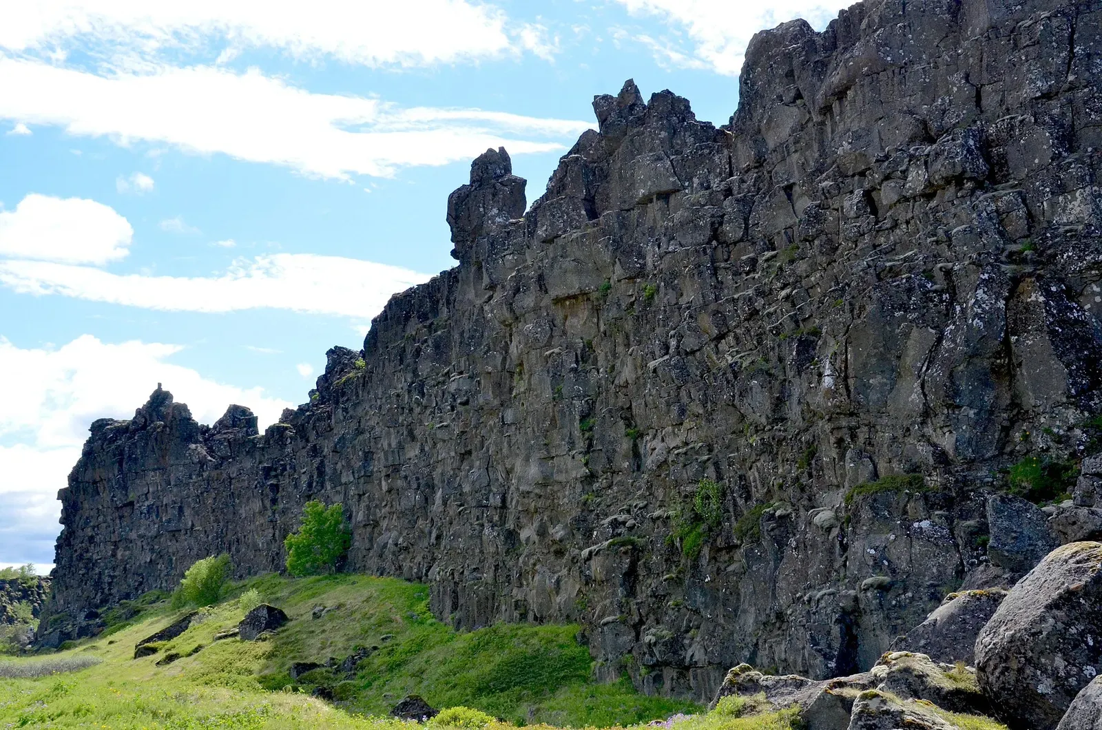 The Almannagjá rift wall rises above green grassland in Þingvellir National Park under a partly clouded sky — Silfra