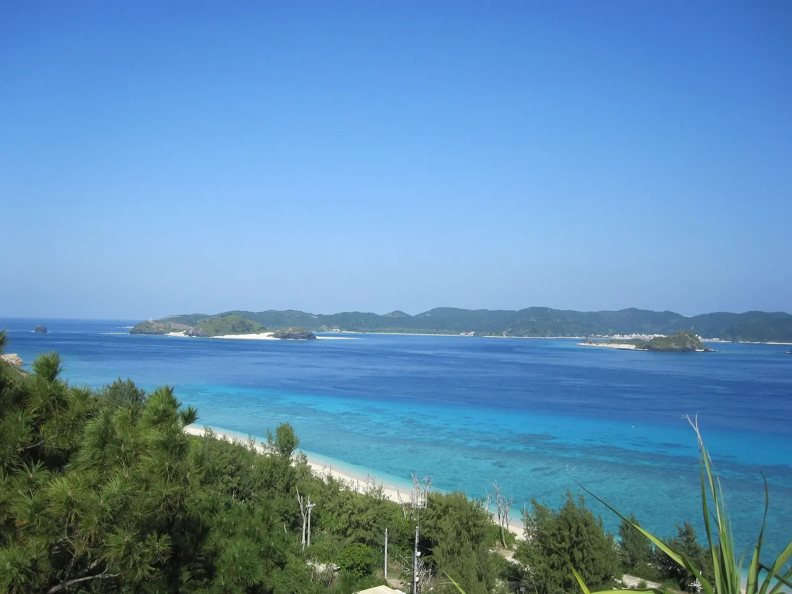 White sand curves into pale turquoise shallows below a green headland on Aka Island — Kerama Islands.