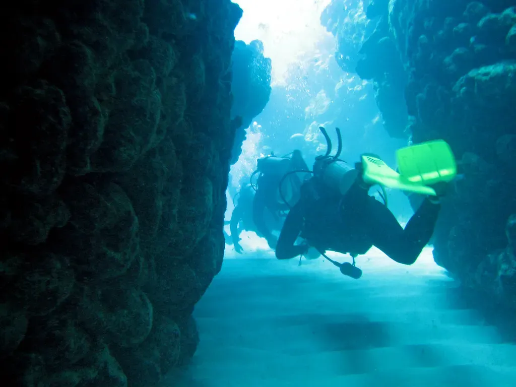 A diver with green fins swims through a narrow coral canyon lit by sunlight from above — Bermuda.