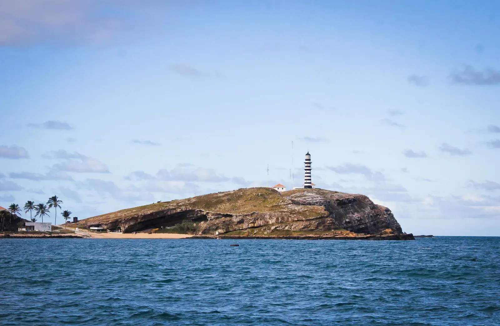The striped lighthouse on Santa Bárbara island stands above a rocky shore and palms, seen from the sea at Abrolhos — Abrolhos Archipelago