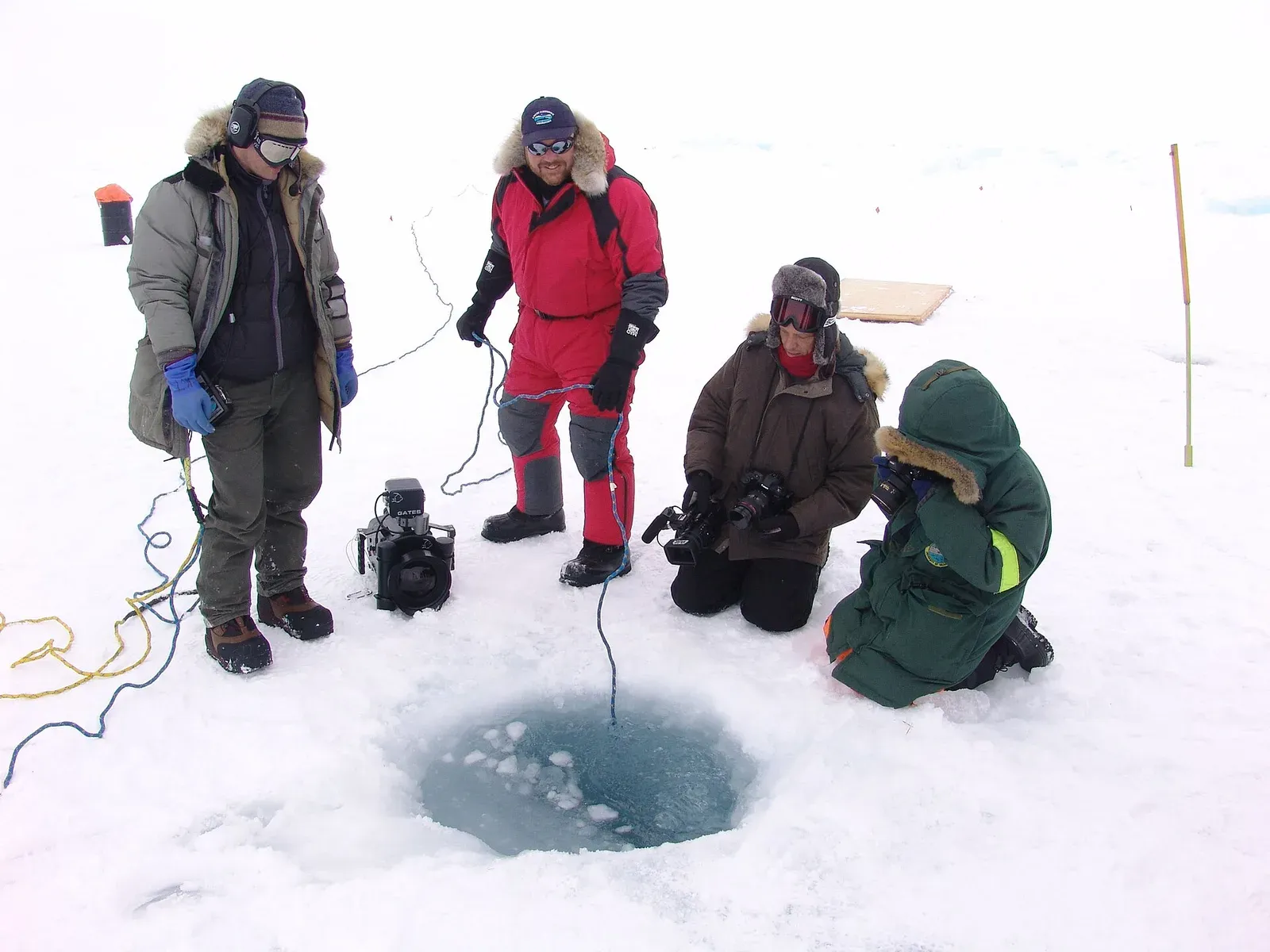 Four people in cold-weather gear gather around a dive hole in Arctic sea ice, tending lines and camera equipment.